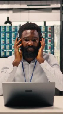 Dikey Video Siyah çalışanı, modern ofiste müzik eşliğinde kulaklık takarak, çalışma alanında neşe ve neşe içinde eğleniyor. Stres atmak için şarkı dinleyen bir adam. Dengeli kurumsal yaşam tarzı.