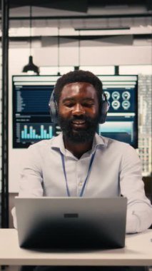 Dikey Video Afrikalı Amerikalı adam iyi ruh hali içinde kulaklıkla müzik dinliyor, bilgisayarla çalışıyor ve stres atışıyla odaklanmış iş analizini birleştiriyor. Ses ve görevler içinde çoklu görev