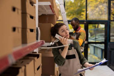 Kadın personel, mal, paket ve küçük teslimat hazırlıklarını yönetirken, sevkiyat, ambalaj ve perakende siparişlerini koordine eden, küçük ölçekli depodaki sabit hatlı bir telefona cevap veriyor..