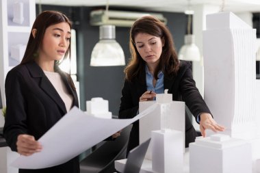 İki kadın mimar beyaz mimari modeli değerlendirir ve basılı inşaat planlarını gözden geçirir. Proje kararları ve uygulama zaman çizelgesi oluşturmada işbirliği yapan profesyonel yaratıcı tasarımcılar.