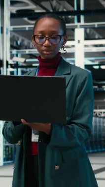 Veri merkezinde yürüyen dikey video programcısı sistemleri izlemek için AR simülasyonu ve dizüstü bilgisayar kullanır. Sunucu merkezindeki Afrikalı Amerikalı kadın optimizasyon ve VFX birleşimi için dizüstü bilgisayar kullanıyor.