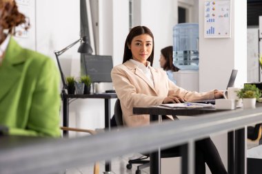 Kendine güveni tam bir bayan yönetici, kameraya odaklanmış ve hazır bir şekilde bakar. Asyalı iş kadını masa başında oturmuş pazarlama firmasının başarısını güvence altına almaya kararlı olduğunu ifade ediyor..