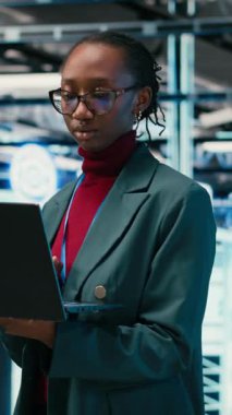 Veri merkezinde yürüyen dikey video programcısı sistemleri izlemek için AR simülasyonu ve dizüstü bilgisayar kullanır. Sunucu merkezindeki Afrikalı Amerikalı kadın optimizasyon ve VFX birleşimi için dizüstü bilgisayar kullanıyor.