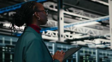 Tablet uygulaması kullanılarak kaynak optimizasyonu için veri merkezi programcısı kod yazıyor. Afrika kökenli Amerikalı bir kadın sunucu merkezine giriyor. Ekipman geliştirmek için programlama metni kullanıyor. Kamera B