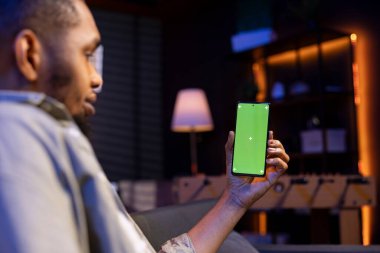 Kanepesinden yeşil ekran kullanan Afrikalı Amerikalı genç adam modern rahat evinin keyfini çıkarıyor. Oturma odasında çalışan rahat bir insan, birden fazla iş yapıyor..