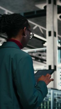 Veri merkezinde kaynak optimizasyonu yapan dikey video yazma kodu, tablet uygulaması ve programcı. Yükseltme ekipmanı, programlama metni ve sunucuda yürüyen Afrikalı Amerikalı kadın.