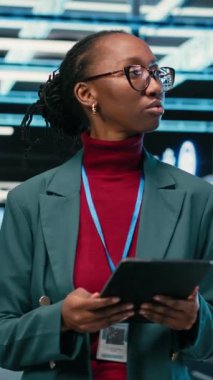 Dikey video mühendisi veri merkezinde, hata kayıtlarını ve hata ayıklama sistemlerini izlemek için tablet ve AR teknolojisini kullanarak yürüyor. Sunucu merkezindeki Afro-Amerikalı kadın mavi kablo şebekesinde tarama yapıyor.