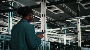 Veri merkezinde kaynak optimizasyonu yapan kod, tablet uygulaması ve programcı yazılıyor. Yükseltme ekipmanları, programlama senaryosu ve sunucu merkezinde yürüyen Afrikalı Amerikalı kadın, kamera A