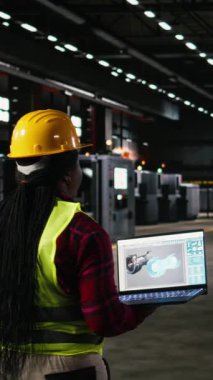 Dikey Video Siyah kadın mühendis, türbin varlık verileri, enjeksiyon sistemleri, dizüstü bilgisayar ekranı. Ağır ekipman, çelik işçiliği ve modern gücü kullanan gelişmiş aletlerle dolu makine dairesi.