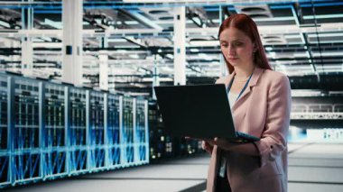 Sunucu çiftliğinde çalışan gülümseyen bir kadının portresi ağ yönetimi viralizasyonu yapıyor. Neşeli veri merkezi kadın teknisyeni laptop ile büyük ölçekli bilişim altyapı süper bilgisayarlarını denetliyor.