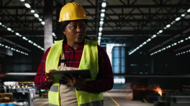 Afrika kökenli Amerikalı kadın personel, bir tablet üzerindeki ağır makine ve iskelet bileşenlerini inceliyor, modern mühendislik fabrikası. Büyük ölçekli üretim hattı tarafından tanımlanan metal çalışma ortamı.