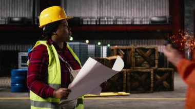 Siyahi kadın işçi, geniş bir sanayi deposunun planlarını ve inşaat çizimlerini inceliyor. Üretim salonunda metal işçiliği, çelik kirişler ve gelişmiş üretim makineleri sergileniyor.