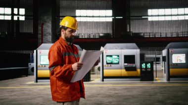 Makine mühendisi modern bir atölyede tasarım ve devre çizimleri üretiyor. Depo ortamı, büyük ölçekli makineler için hassas ölçümleri vurguluyor.