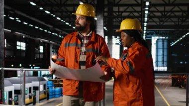 Çok ırklı iş arkadaşları, teknik planlar, detaylı çizimler, ağır sanayi santrali, üretim planları, çalışma ortamı, ağır makineler ve üretimi optimize etmeye odaklı personel.