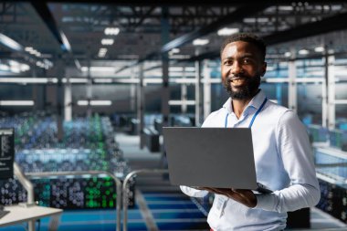 Sunucu odasındaki kendinden emin mühendisin portresi endüstriyel platformdaki yapay zeka görselleştirmesini inceleyerek, derin öğrenme yazılımı gelişimini izliyor. Genç adam operasyonel güvenilirliğini koruyor.