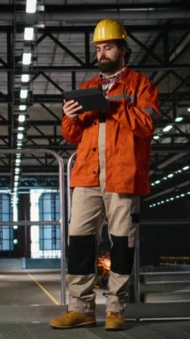 Dikey Video Mühendisliği çalışanı ağır sanayi üretim hattını tabletle, makine deposuyla denetliyor. Metal ve endüstriyel ekipmanlarla büyük ölçekli üretim süreci, bitki