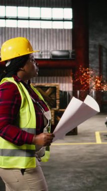 Dikey Video Siyah kadın çalışan, geniş bir sanayi deposundaki planları ve inşaat çizimlerini analiz ediyor. Üretim salonunda metal işçiliği, çelik kirişler ve gelişmiş makineler sergileniyor