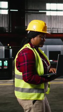 Dikey Video Kadın Mühendis Endüstri Salonu boyunca makineleri kontrol ederek, Dizüstü bilgisayardaki üretim akışını ve imalat operasyonlarını izliyor. Metalik kirişlerle dolu ağır sanayi ortamı.