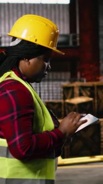 Dikey Video Siyah kadın, endüstriyel mühendis, üretim hattını denetleyen, tablet üzerinde ağır sanayi depolama tesisinde teftiş yapan. Kaynak istasyonları ve demir fabrikaları olan büyük ölçekli bir fabrika.