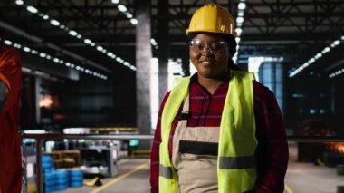 Portre, siyah kadın, güvenlik gözlükleri ve miğfer, üretim salonu mühendisliği, üretim sürecinde büyük çaplı çelik işçiliği. Ağır makine faaliyetlerini denetleyen kadın mühendis..