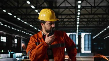 Mühendis, ağır endüstri deposunda sabit şapka takmış taşınabilir telsizle konuşuyor. Kablosuz radyo aygıtı, büyük ölçekli makine salonu boyunca temiz iletişim ve koordinasyon sağlar.