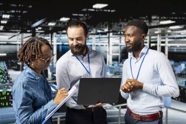 Multiethnic employees team collaborating on maintenance for server rigs and handling AI processing tasks. Data center engineering team supporting machine learning for software development.