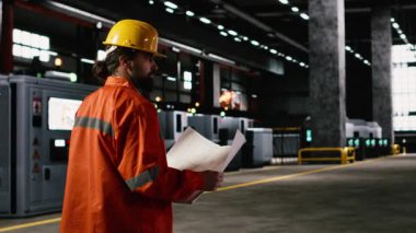 Sert şapkalı usta mühendis, ağır sanayi atölyesindeki teknik planları ve çizimleri inceliyor. Çelik işçiliğinin yanındaki sanayi bölgesinde yürüyen ve kaynak makinelerinden kıvılcımlar saçan bir adam..