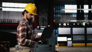 Yetişkin mühendis, büyük ölçekli bir depoda üretim hattını değerlendiriyor, miğfer takıyor ve mühendislik fabrikasından geçiyor. Ağır sanayi atmosferi için büyük makineler ve çelik işçiliği.
