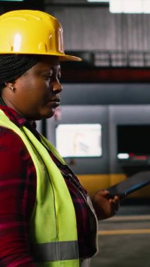 Dikey Video Kadın Mühendisi, üretim salonu boyunca endüstriyel işletim istasyonlarını tabletle denetliyor. Depo tesisinde ağır endüstriyel metaller var. Kıvılcımlar saçan mühendislik fabrikası