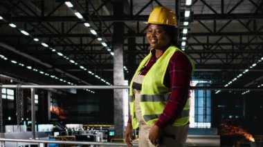 Ağır endüstri mühendisliği fabrikasında çalışan, kask takan ve büyük ölçekli üretim hattında metal işleriyle uğraşan siyahi bir kadının portresi. Fabrikada üretim süreci.