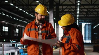 Çok ırklı personel, ağır sanayi makinelerinin bulunduğu geniş bir depoda teknik operasyonların planlarını ve çizimlerini inceliyor. Büyük ölçekli üretim hattını desteklemek.