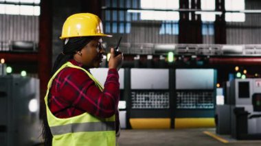 Zenci kadın mühendis taşınabilir telsiz cihazına konuşuyor, depoda yürüyor ve ağır ekipmanları izliyor. Büyük ölçekli sanayi tesisi üretim hattını ön plana çıkardı.