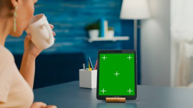 Masanın üzerinde duran yeşil ekran krom anahtarlı tablet bilgisayara bakarken bir fincan kahve içen kadın. İnternet iletişim projesi için izole edilmiş aygıt kullanan serbest çalışan