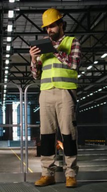 Miğfer takan ve tablet üzerinde üretim hattını kontrol eden dikey video mühendisi devasa makinelerle dolu endüstriyel alanda yürüyor. Depo ortamında ağır sanayi