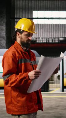 Dikey Video Mekanik Mühendisi, modern bir atölyede üretim planları ve devre çizimleri inceliyor. Depo ortamı, büyük ölçekli makineler için hassas ölçümleri vurguluyor