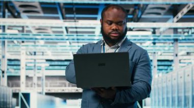 Sunucu merkezindeki mutlu programcı altyapı makinelerinin bakım işlerini yapmak için dizüstü bilgisayar kullanıyor. Modern yüksek teknoloji veri merkezindeki neşeli adam sunucu ayarlarını not defteri, kamera B ile yapılandırıyor.