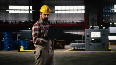 Yetişkin mühendis, büyük ölçekli bir depoda üretim hattını değerlendiriyor, miğfer takıyor ve mühendislik fabrikasından geçiyor. Ağır sanayi atmosferi için büyük makineler ve çelik işçiliği.
