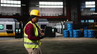 Siyahi kadın mühendis, endüstriyel salonda makineleri kontrol ediyor. Üretim akışını ve imalat operasyonlarını dizüstü bilgisayardan izliyor. Metalik kirişlerle dolu ağır sanayi ortamı.