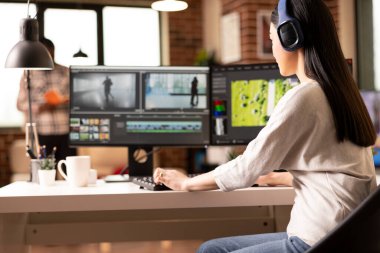 Kablosuz kulaklıklı serbest video editörü tuğla duvar dairesinde sinematik içerik yaratıyor. İkili masaüstü monitörleri ve görsel hikaye anlatma ve VFX yazılımı olan bir kadın çalışma istasyonunda oturuyor.