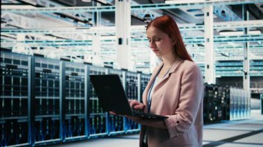 Sunucu odasındaki mutlu kadın, altyapının işlevsel etkinliğini arttırmak için not defteri kullanıyor. Veri merkezindeki neşeli kadın mühendis, donanım ölçümlerini, kamera A 'yı izlemek için laptop kullanıyor.