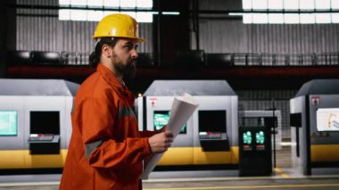 Makine mühendisi modern bir atölyede tasarım ve devre çizimleri üretiyor. Depo ortamı, büyük ölçekli makineler için hassas ölçümleri vurguluyor.