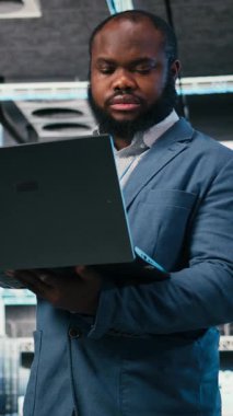 Laptop kullanarak, ağ yönetimi viralizasyonu yaparak sunucu çiftliğinde dikey video yazılım geliştirici. Veri merkezindeki Afrikalı Amerikalı adam bilgisayarların bilgisayar altyapısını denetliyor.