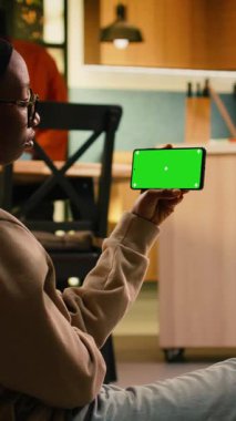 Dikey video izole krom anahtar, akıllı telefon ekranı, yerde dinlenen siyah kadın, sıcak gün ışığında evde oturuyor. Koridorda telefonu tutan genç bir yetişkin, günlük ev hayatı...