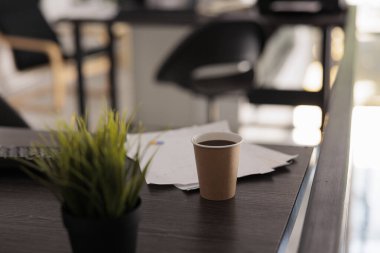 Close up view of disposable coffee cup on wooden desk in early morning office. Papers with work reports and indoor plant, background out of focus shot of desks in workplace.