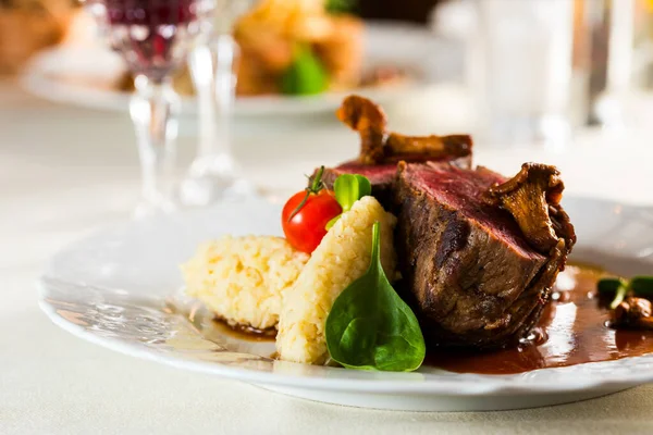 a side dish of porridge and two medium fried steaks with basil decorations of tomatoes and mushrooms on a platter in a restaurant