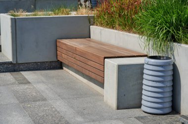 Bir dizi koltuk ve katmanlı beton elementlerden oluşan şehir manzarası tasarımı. Ön planda açık gri renkli silindirik atık konteynırı var. Ahşap kafes tezgahı ve katları beton desenli gri taştan yapılmıştır..