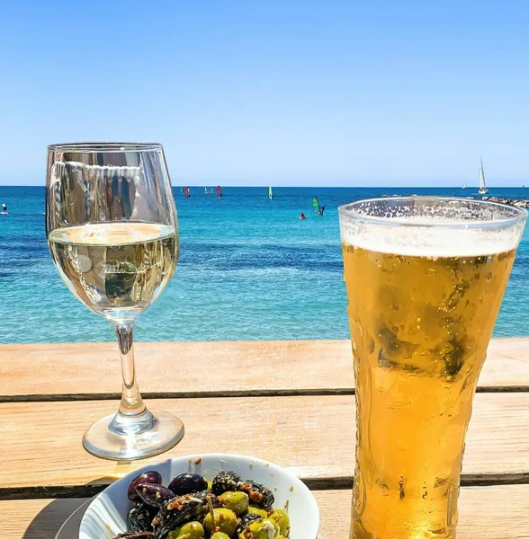 Wooden table with light beer in the mug , white wine in the glass and olives in white plate at the beach on blue sea and sky background.