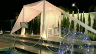 Yahudi gelenekleri, açık hava düğün töreni. Düğün örtüsü (chuppah, hupa) akşam gökyüzünün arka planında beyaz kumaştan yapılmış, palmiyeler ve balonlarla süslenmiş yapay bir rezervuar. 4K