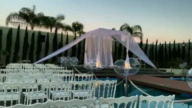Yahudi gelenekleri, açık hava düğün töreni. Düğün örtüsü (chuppah, hupa) akşam gökyüzünün arka planında beyaz kumaştan yapılmış, palmiyeler ve balonlarla süslenmiş yapay bir rezervuar. 4K