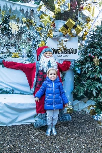Leuk blond meisje, 4 jaar oud, met roze hoepel in haar haren en blauwe jas in de buurt van Santa Claus — Stockfoto